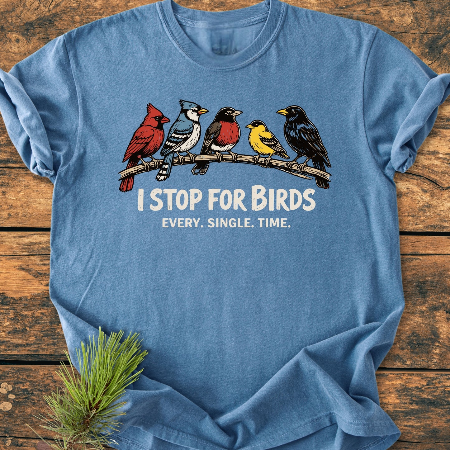 I Stop For Birds - Vintage Wash Tee features five songbirds on a branch with slogan text, laid on wood with a pine sprig, highlighting its relaxed, soft-washed design and durable stitching.
