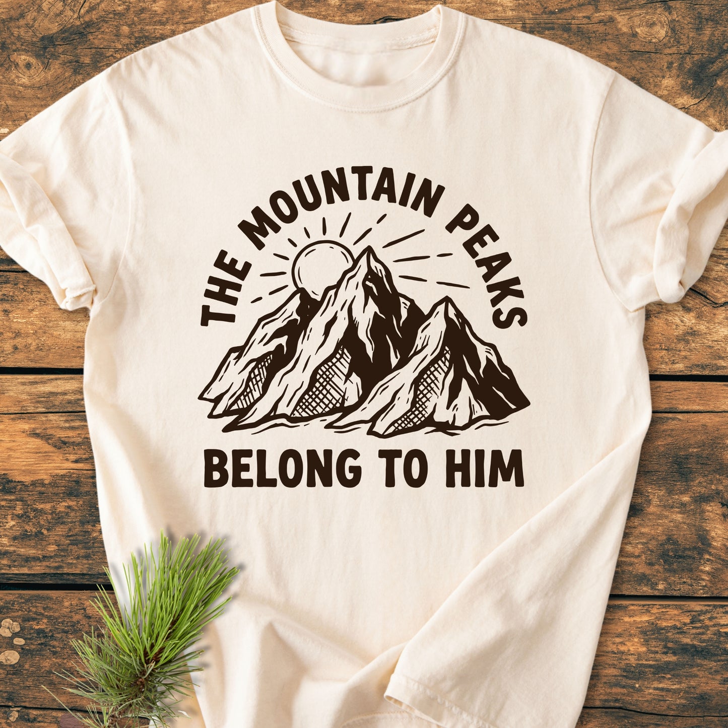 The Mountain Peaks Belong To Him - Vintage Wash Tee features a line-drawn mountain and sun graphic with bold text, laid flat on wood, accentuated by a pine sprig, showcasing its relaxed, durable design.