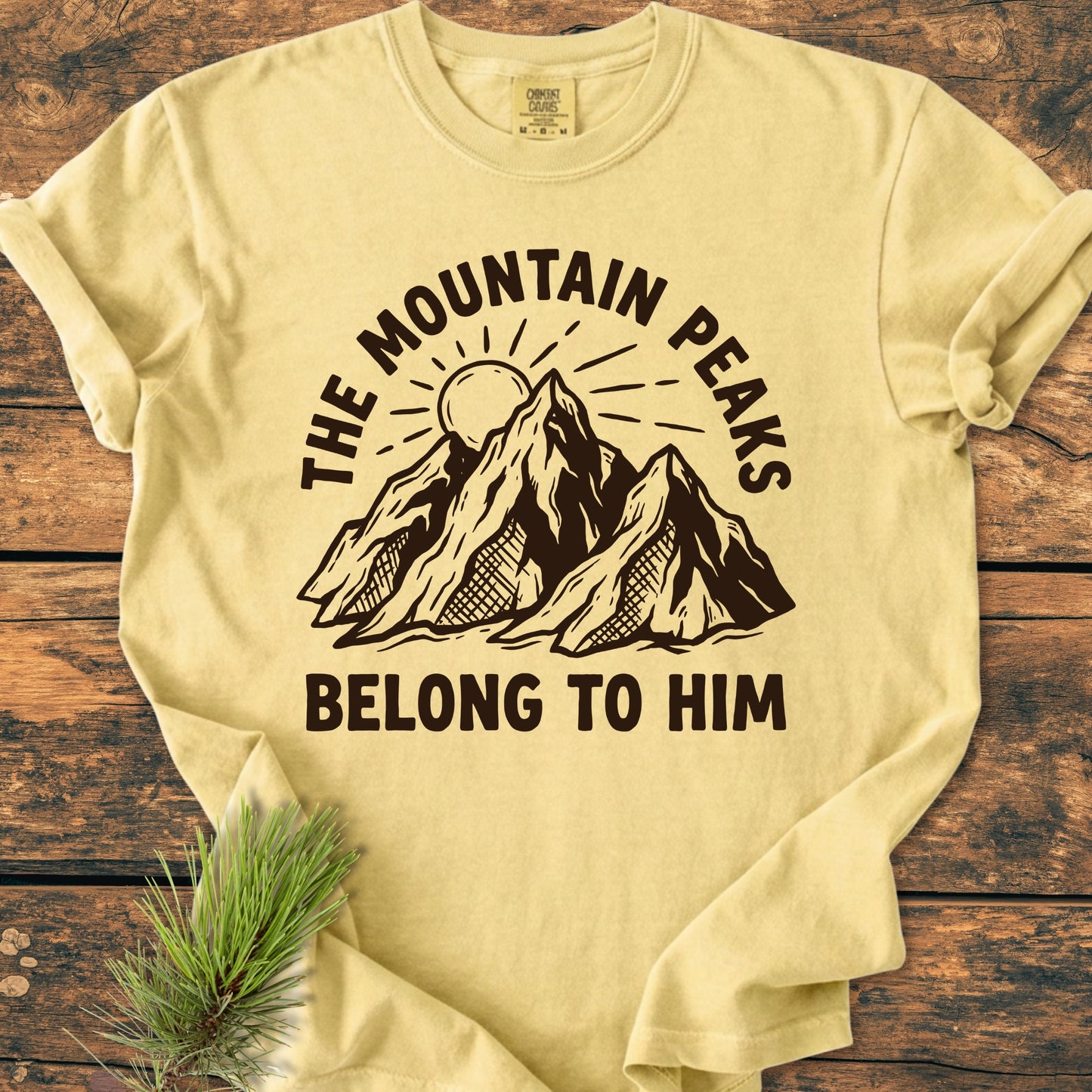 The Mountain Peaks Belong To Him - Vintage Wash Tee features a stylized mountain print with a sun, laid flat on wood, accented by a pine sprig, highlighting its natural theme.