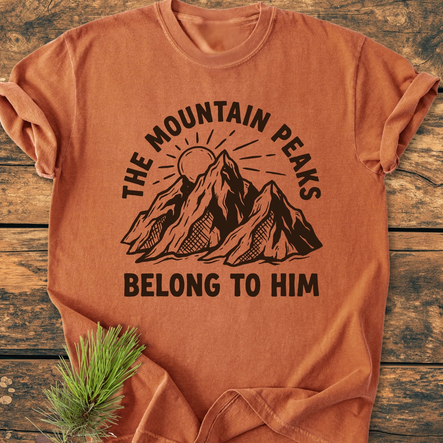 The Mountain Peaks Belong To Him - Vintage Wash Tee features a bold mountain graphic and slogan, laid on wood with a pine sprig, showcasing its rustic, relaxed style.