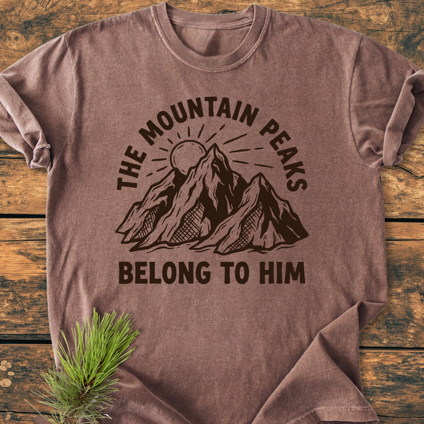 The Mountain Peaks Belong To Him - Vintage Wash Tee features a mountain and sun graphic on heathered brown fabric. Laid on wood, it includes a pine sprig accent for an outdoorsy touch.