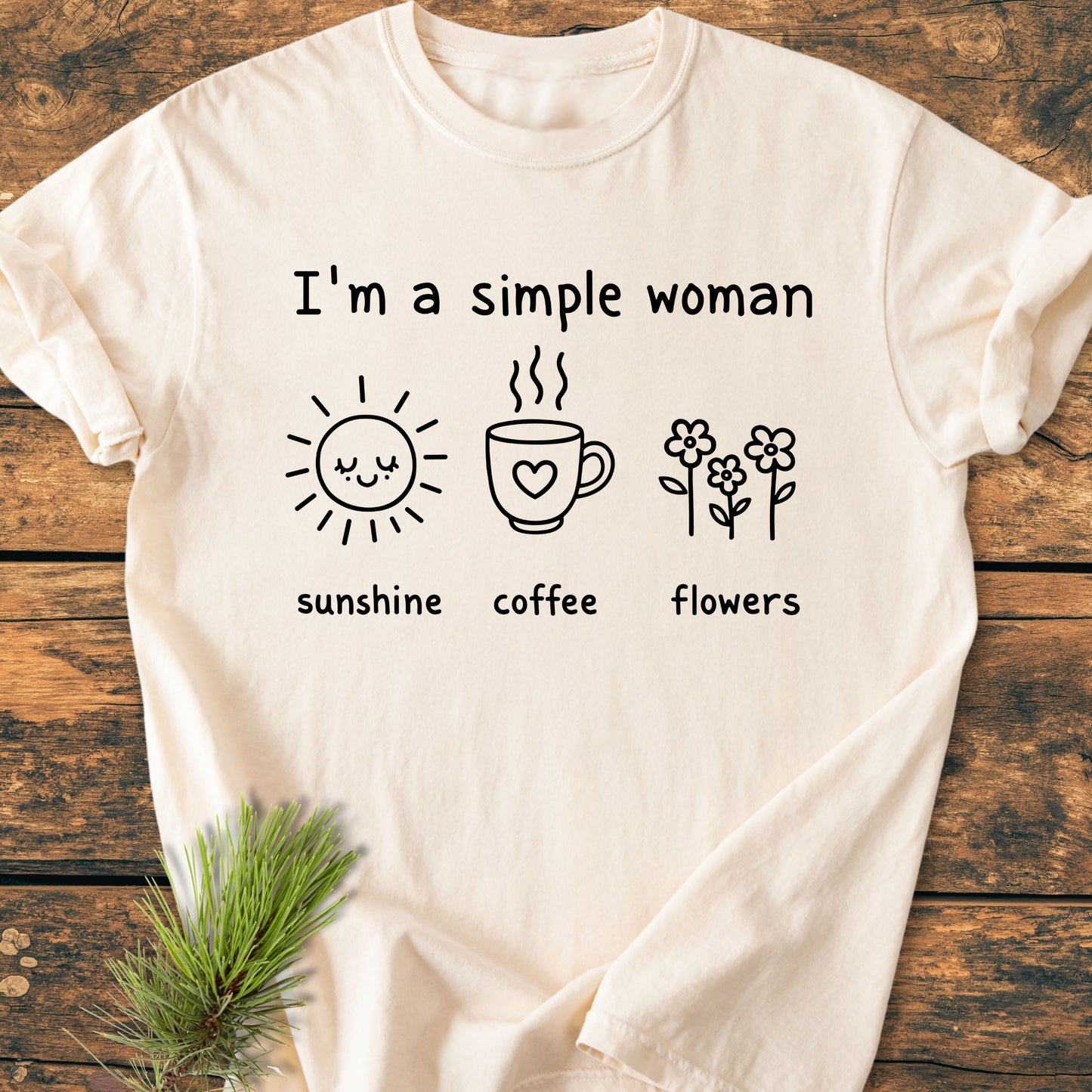 I'm A Simple Woman. Sunshine Coffee Flowers - Vintage Wash Tee laid on wood, featuring a black hand-drawn design with sunshine, coffee, and flowers icons. Pine sprig decorates beside the t-shirt.