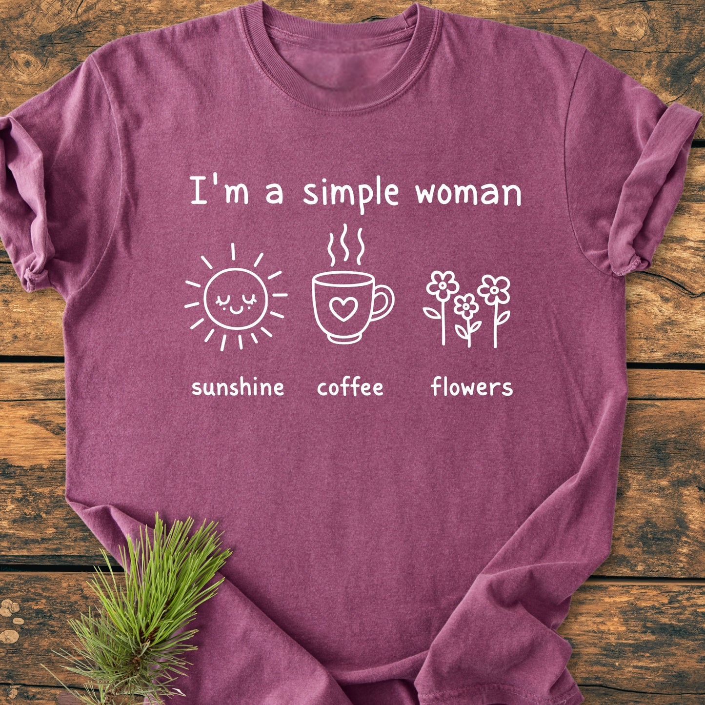 I'm A Simple Woman. Sunshine Coffee Flowers - Vintage Wash Tee in plum, featuring white illustrations of a sun, coffee mug, and flowers, laid on wood with a pine sprig beside it.