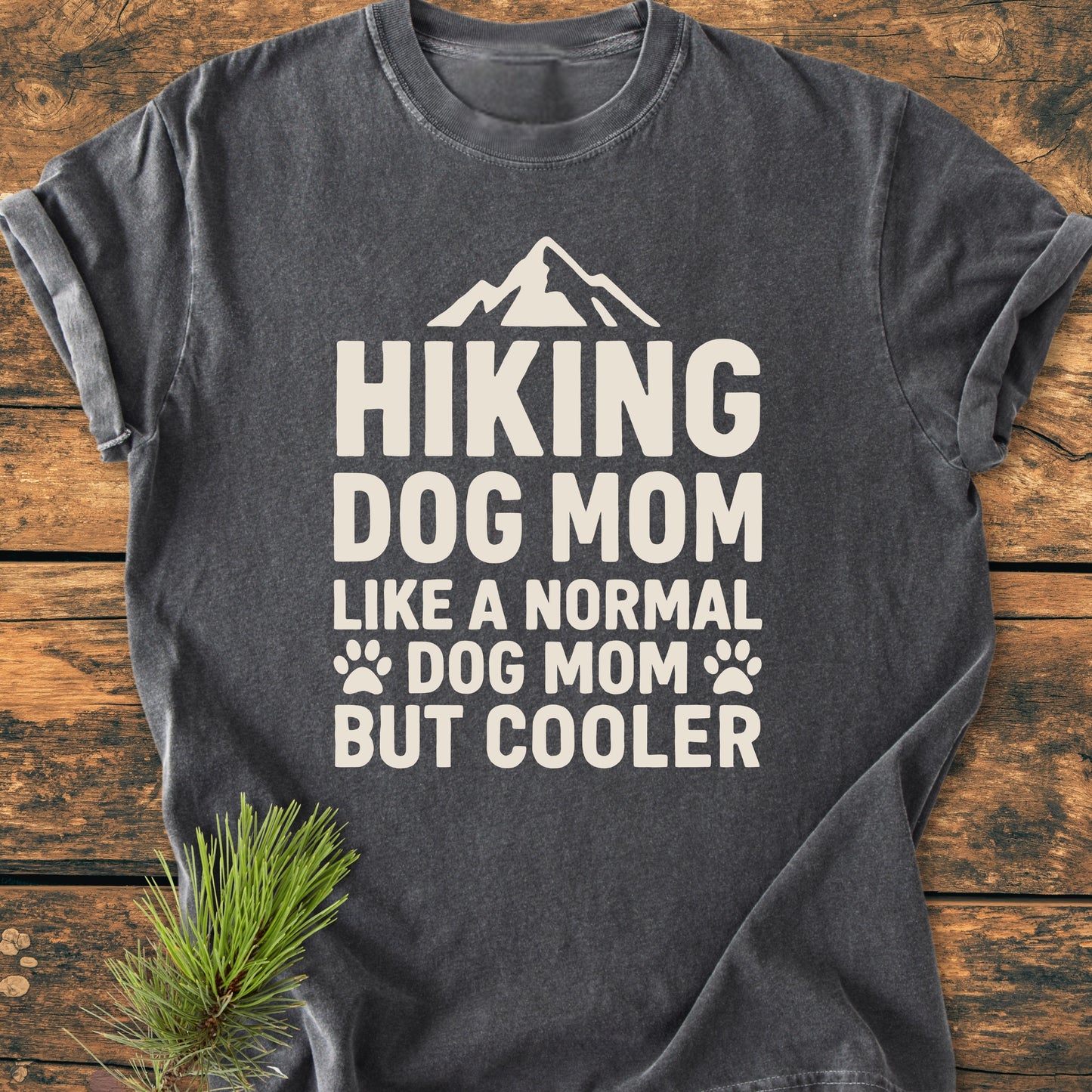 Charcoal-gray Hiking Dog Mom - Vintage Wash Tee with cream-white mountain design, laid flat on wooden surface with pine sprig. Cozy, relaxed fit; perfect for daily wear.