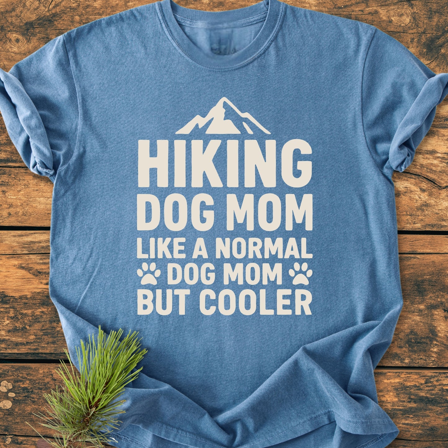 Heather-blue Hiking Dog Mom - Vintage Wash Tee with a white mountain silhouette and playful text, displayed on a wooden surface, emphasizing its cozy, relaxed fit and durable construction.