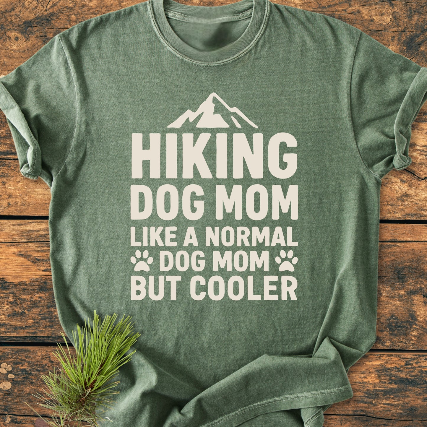 Hiking Dog Mom - Vintage Wash Tee in forest green, featuring a mountain graphic and playful text, laid on wooden surface with a pine sprig, showcasing its outdoorsy, relaxed style.