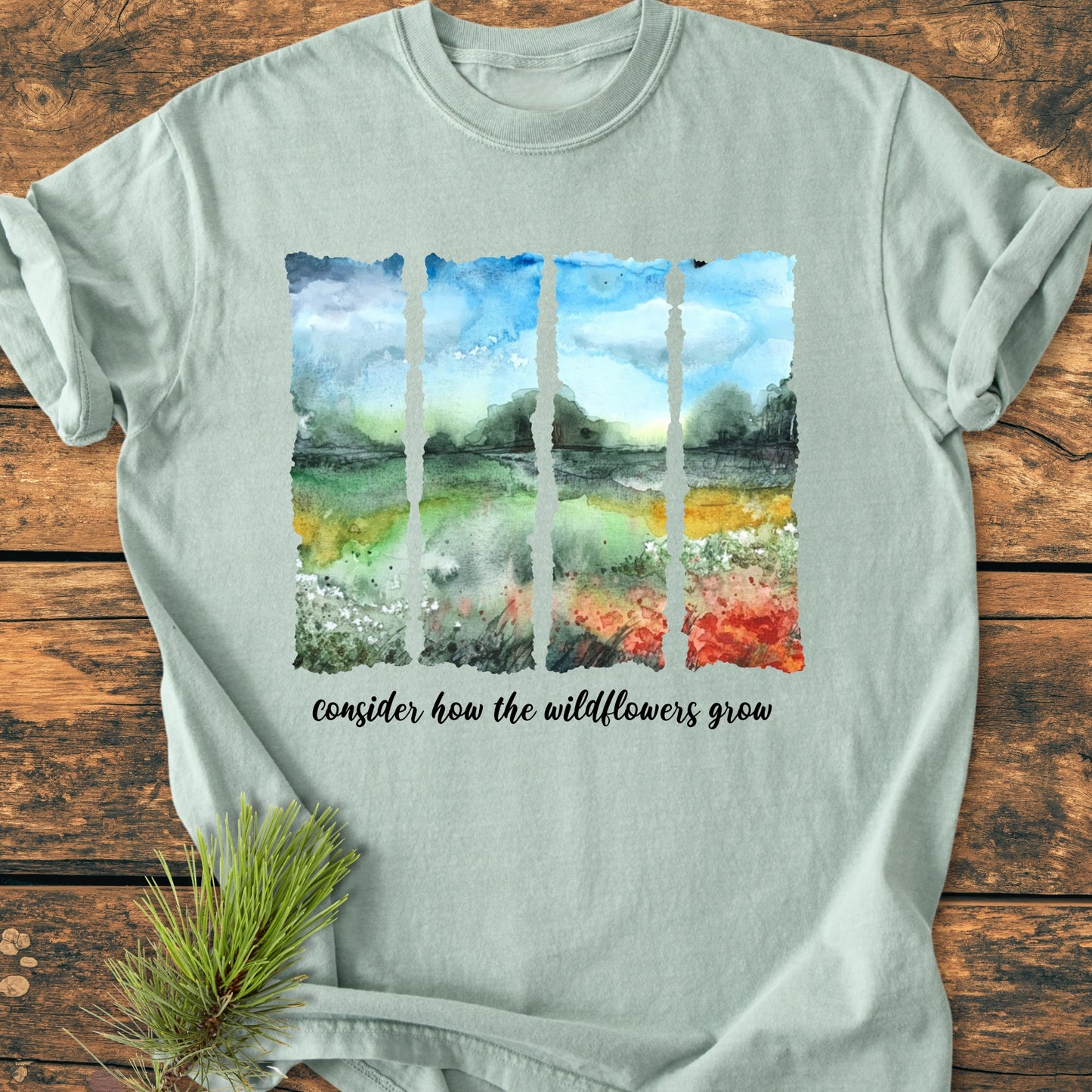 Consider How The Wildflowers Grow - Vintage Heavyweight Tee featuring a watercolor sky and wildflower scene on pale-green fabric, paired with the phrase in black script, lying on a wooden surface.