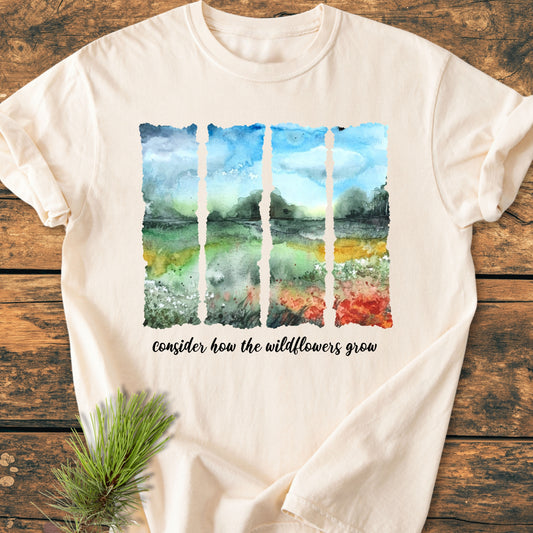 Consider How The Wildflowers Grow - Vintage Heavyweight Tee, featuring a watercolor-style landscape in five vertical panels and a cursive inscription, laid on wood with a pine sprig beside it.