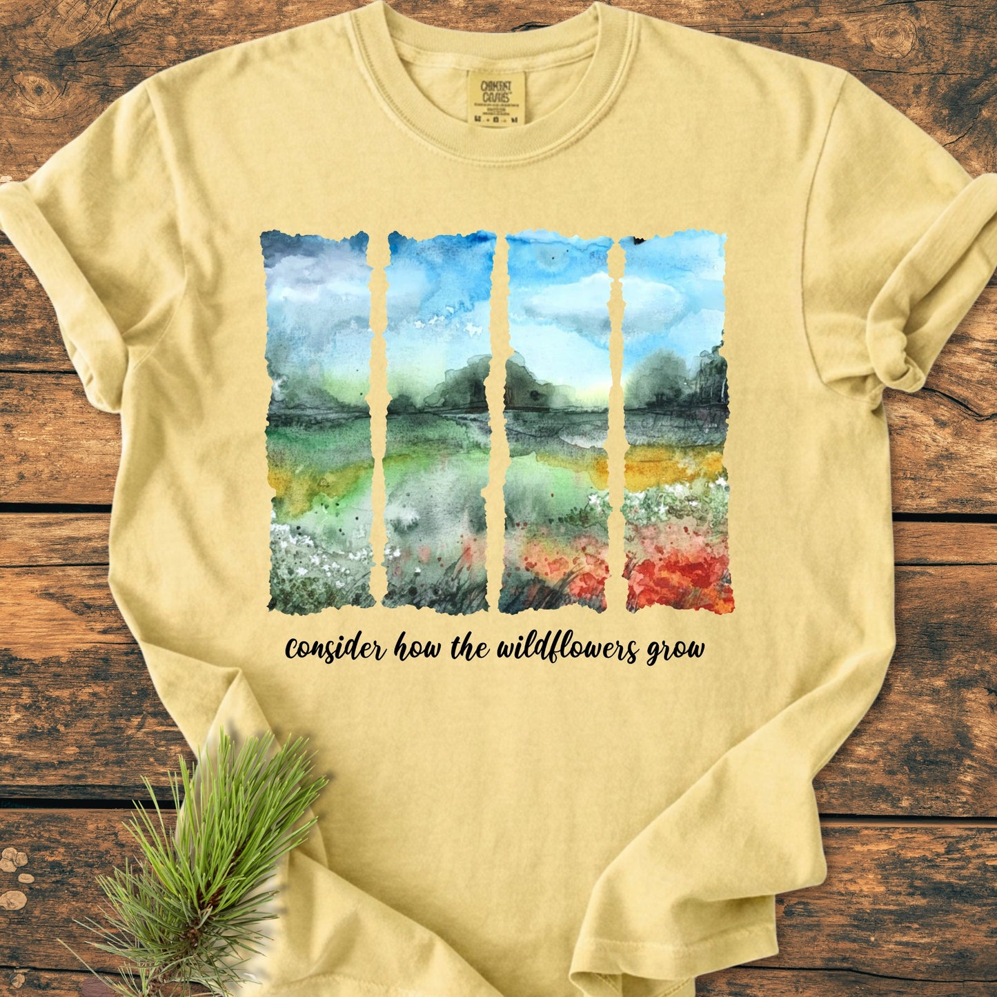 Consider How The Wildflowers Grow - Vintage Heavyweight Tee features a watercolor landscape print on light-yellow fabric, with a pine twig nearby, emphasizing its natural and artistic design.
