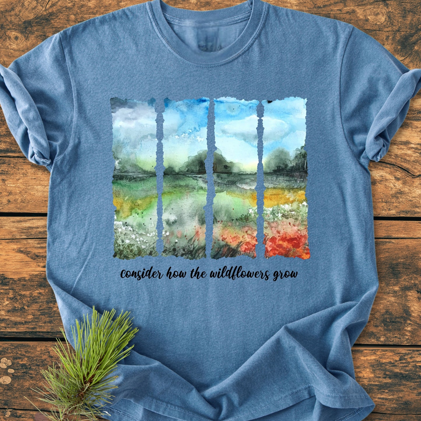 Consider How The Wildflowers Grow - Vintage Heavyweight Tee: Heather-blue T-shirt with a watercolor landscape and hand-lettered phrase, laid on rustic wood, accented by pine sprigs.
