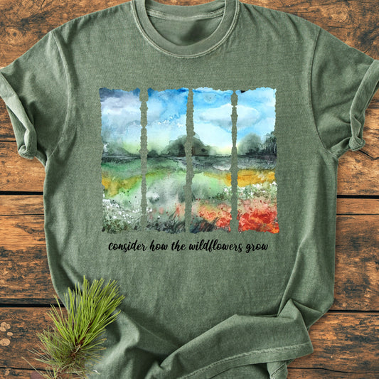 Consider How The Wildflowers Grow - Vintage Heavyweight Tee features a watercolor-style landscape with wildflowers. Laid on rustic wood, it's styled with a pine branch, showcasing its relaxed, durable design.