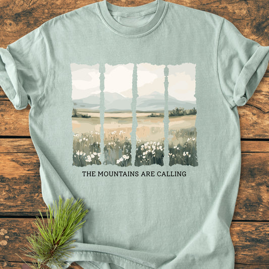 The Mountains Are Calling - Vintage Heavyweight Tee features a muted landscape graphic with wildflowers and mountains, and the phrase “THE MOUNTAINS ARE CALLING,” on a pale-green, short-sleeve T-shirt.