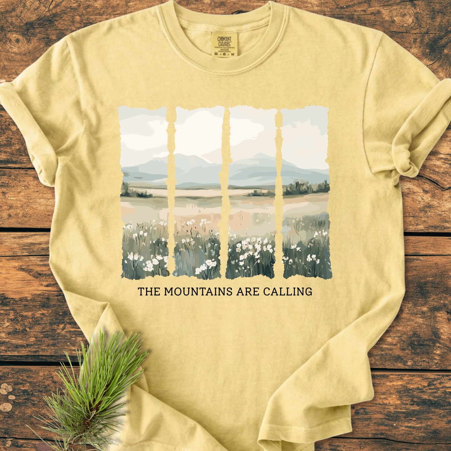 The Mountains Are Calling - Vintage Heavyweight Tee features a painterly mountain scene and slogan on a soft-yellow crewneck, displayed on a rustic wooden surface with a pine sprig nearby.
