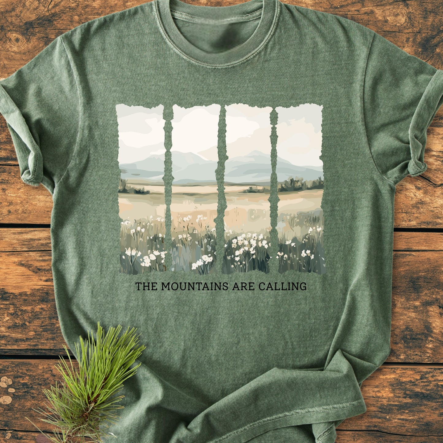 The Mountains Are Calling - Vintage Heavyweight Tee features a wildflower meadow print with distant mountains, laid on wood. A pine sprig accompanies this relaxed, durable, 100% cotton garment.