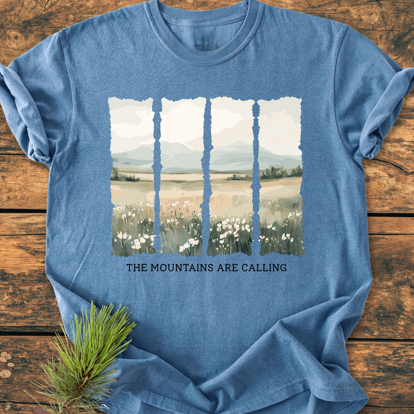 The Mountains Are Calling - Vintage Heavyweight Tee features five torn-edge panels of pastel landscapes on a heather-blue T-shirt, styled on a wooden surface with a pine sprig.