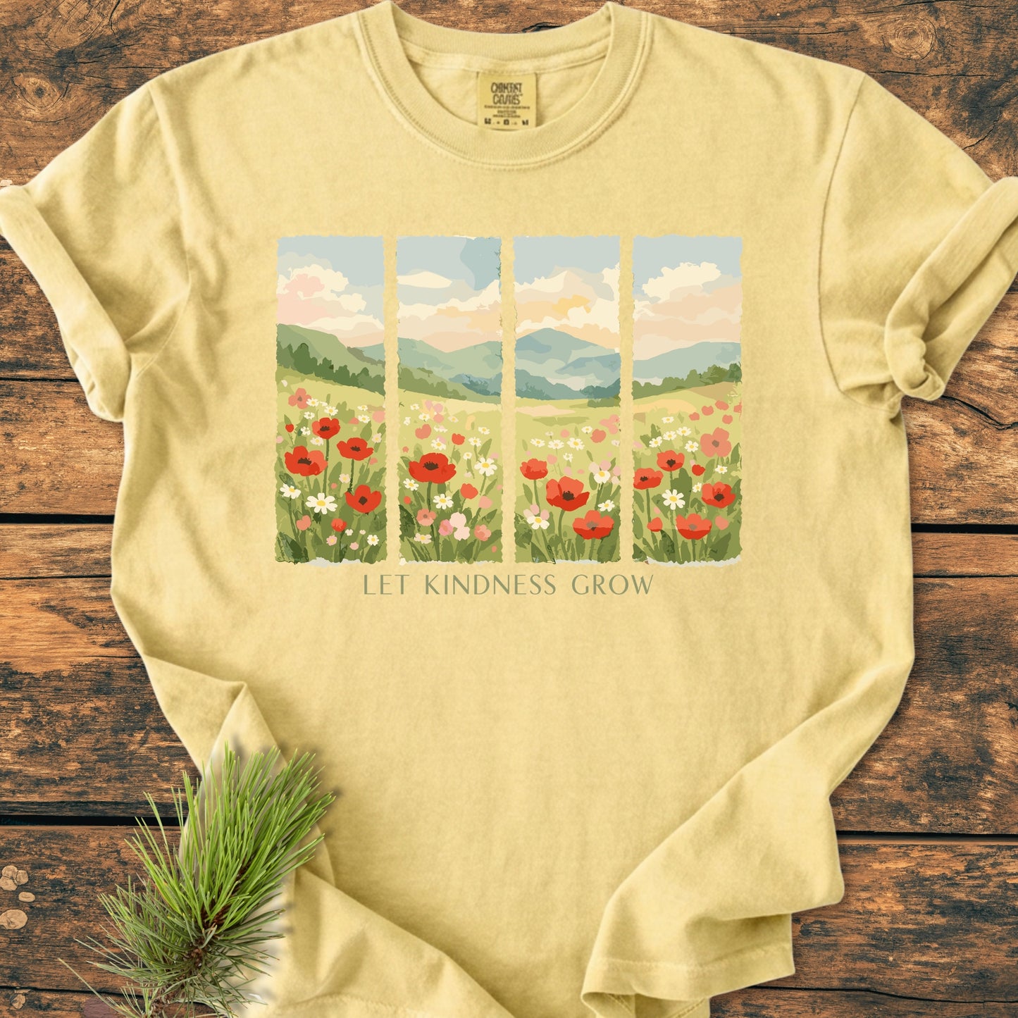 Let Kindness Grow - Vintage Heavyweight Tee features a pastoral scene with wildflowers and slogan, LET KINDNESS GROW, on pale-yellow fabric, laid flat with pine sprig accent.