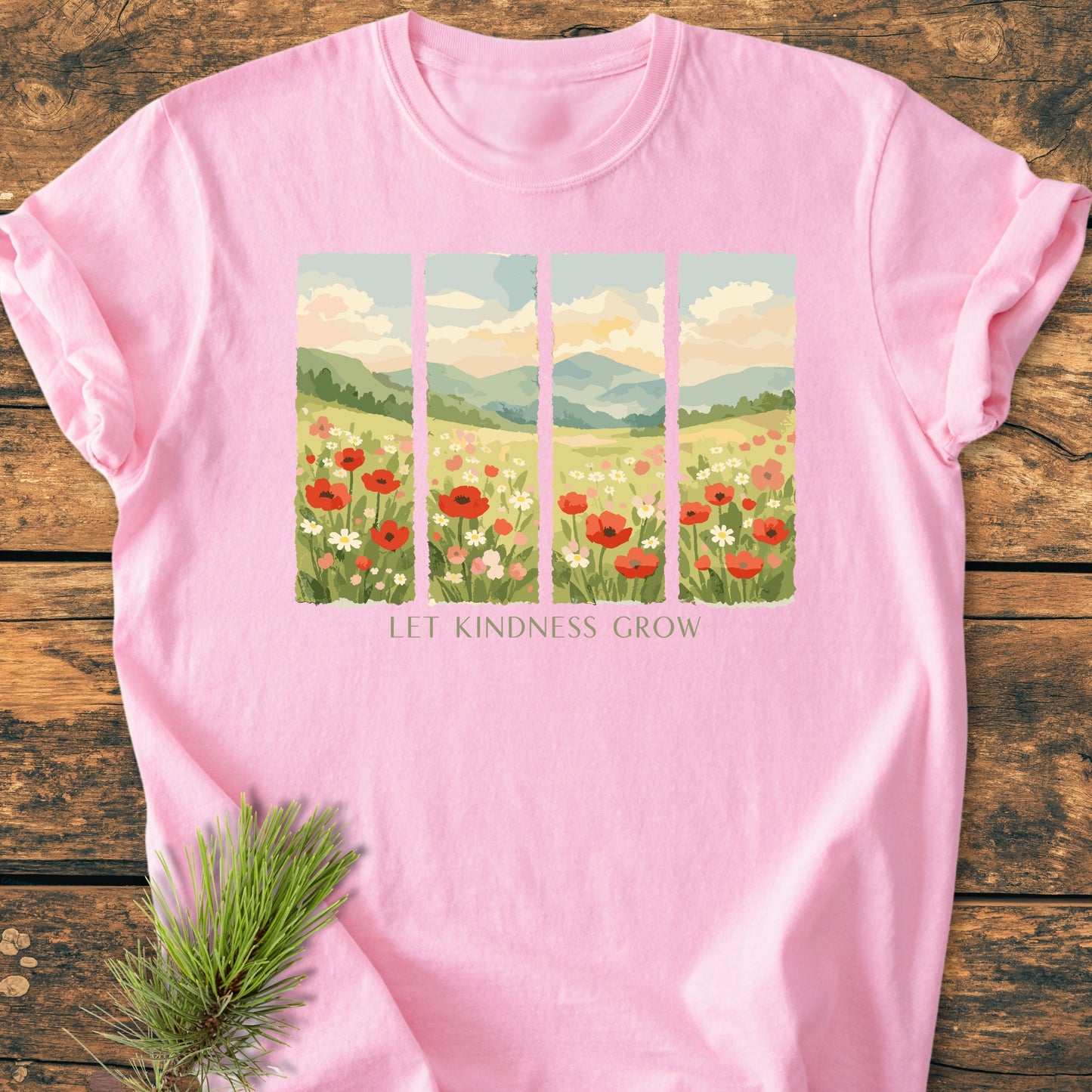 Let Kindness Grow - Vintage Heavyweight Tee features a painterly landscape of hills and flowers with LET KINDNESS GROW slogan on a soft pink T-shirt, laid on rustic wood with a pine sprig.