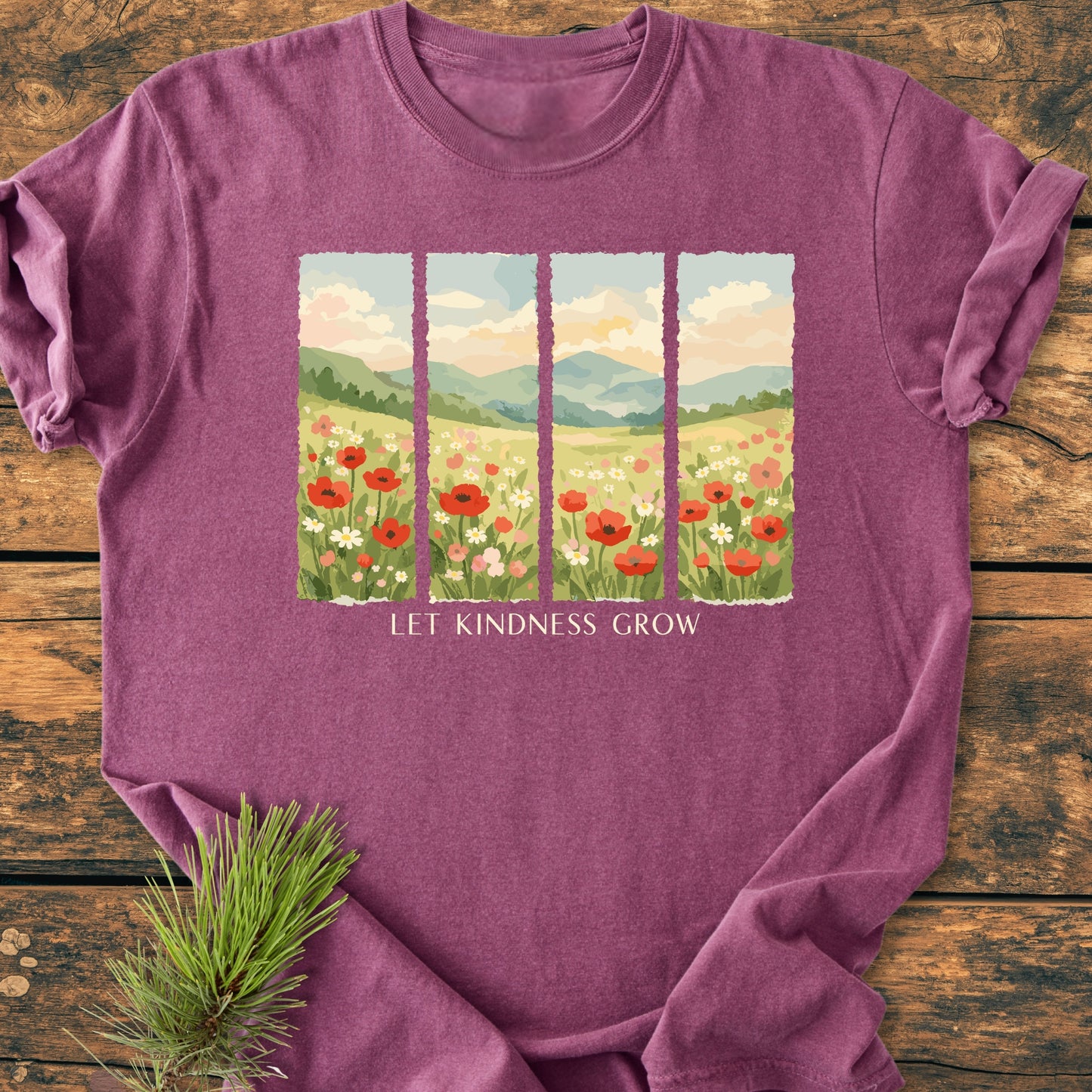 Let Kindness Grow - Vintage Heavyweight Tee with meadow graphic and slogan, laid flat on a wooden surface, exuding casual, nature-inspired styling with an evergreen sprig beside it.
