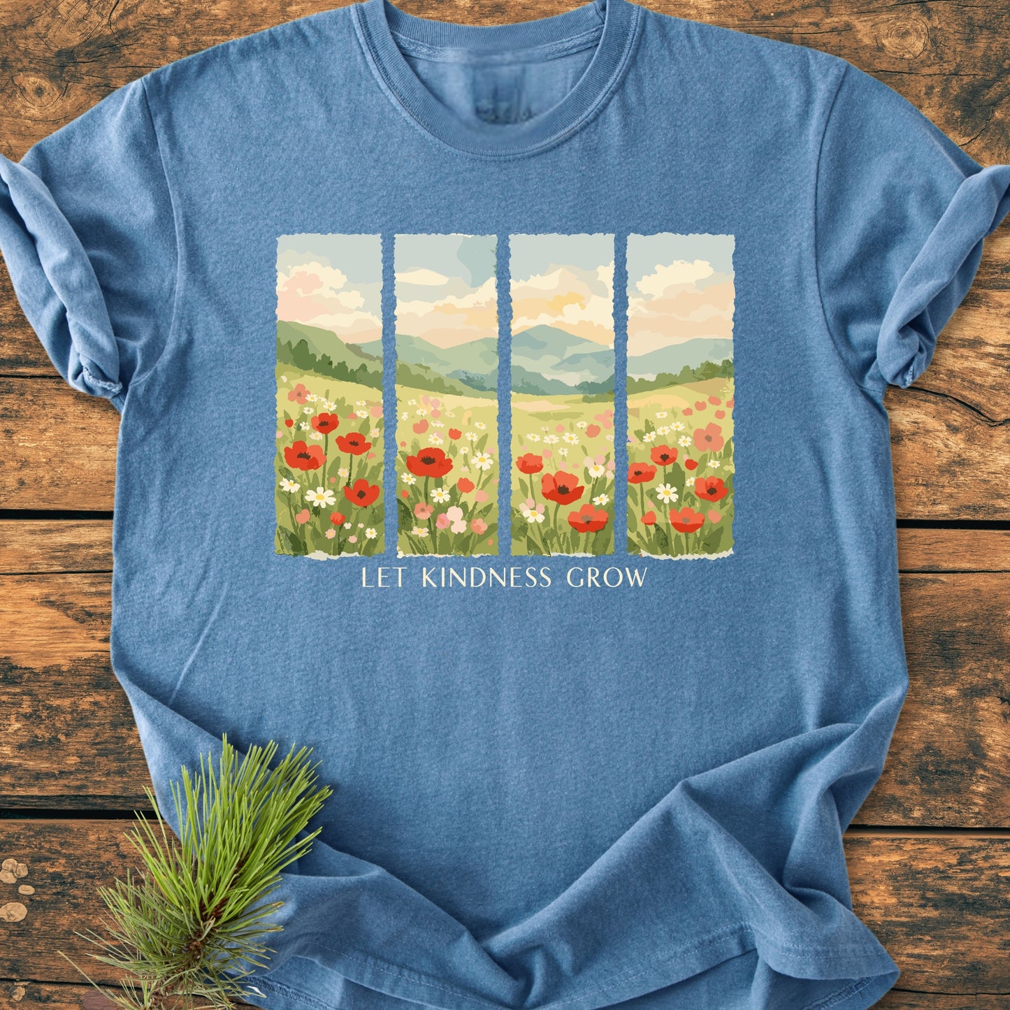 Let Kindness Grow - Vintage Heavyweight Tee featuring a sunlit meadow graphic and slogan, laid flat on rustic wood with a pine sprig accentuating its natural, outdoorsy appeal.