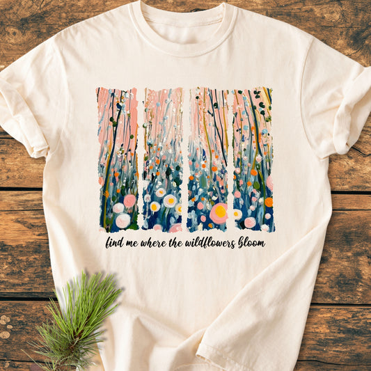 Find Me Where The Wildflowers Bloom - Vintage Heavyweight Tee features a painterly wildflower design and script on a light-cream T-shirt, laid on rustic wood with a pine sprig beside it.