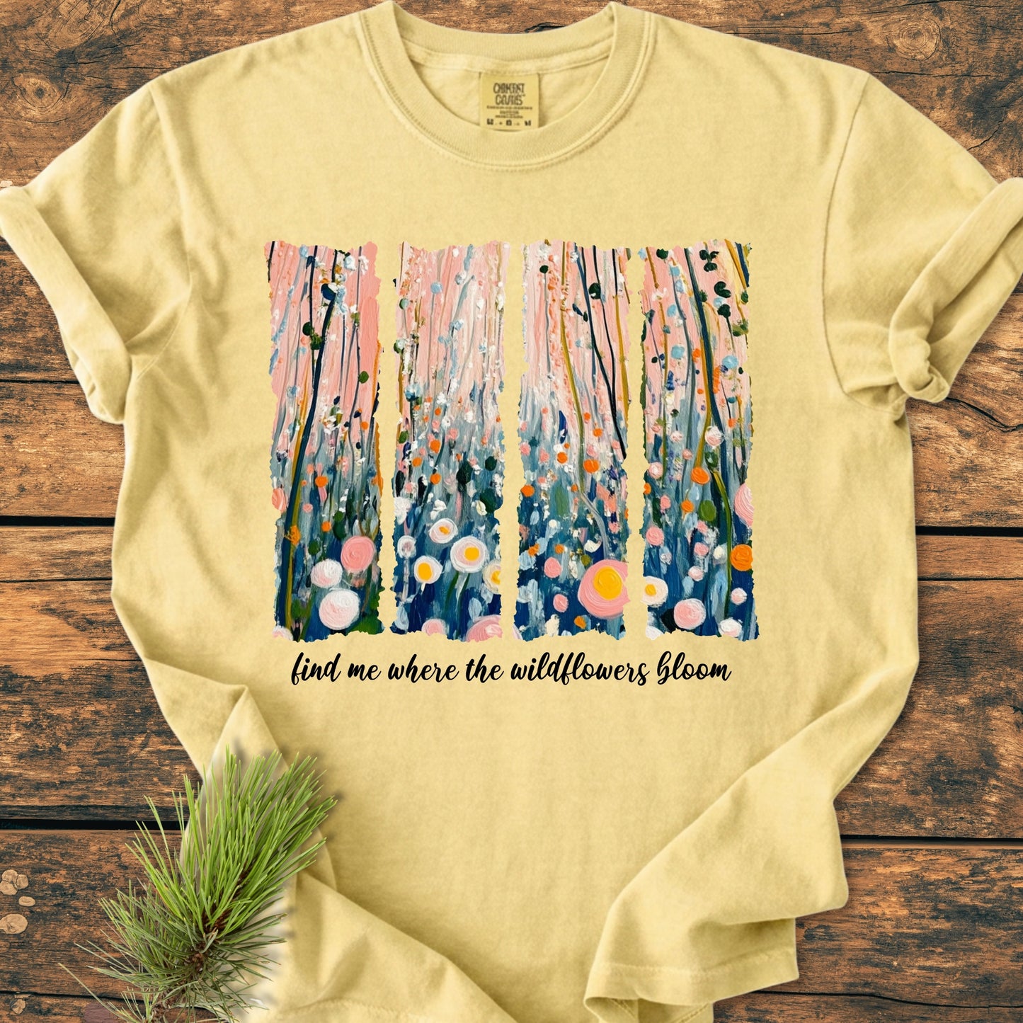 Find Me Where The Wildflowers Bloom - Vintage Heavyweight Tee features an abstract wildflower print in vibrant colors on a pale-yellow, short-sleeve T-shirt, laid on a rustic wood surface.