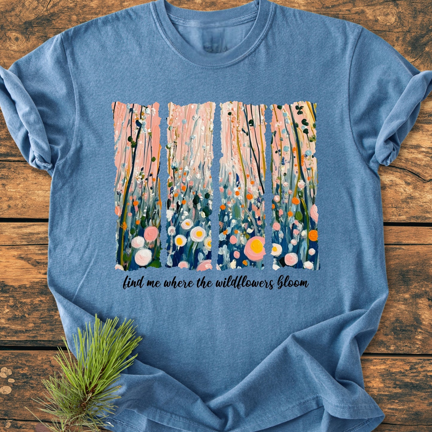 Find Me Where The Wildflowers Bloom - Vintage Heavyweight Tee features an abstract wildflower painting with cursive text on a heather-blue T-shirt, laid flat with a pine sprig accent.