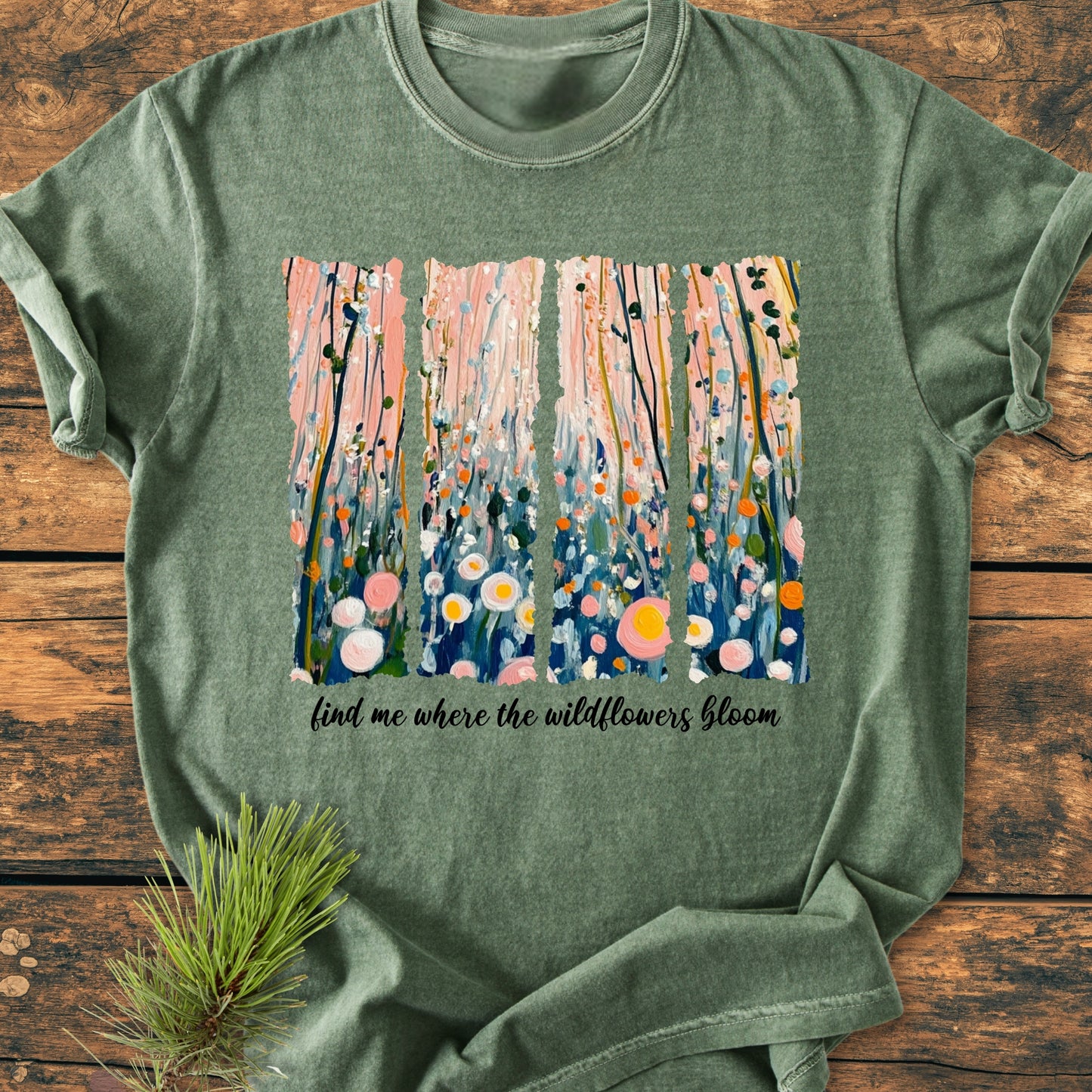 Find Me Where The Wildflowers Bloom - Vintage Heavyweight Tee features a wildflower scene and script on heather-green fabric, laid on wood with a pine sprig accentuating the cozy, relaxed-fit design.