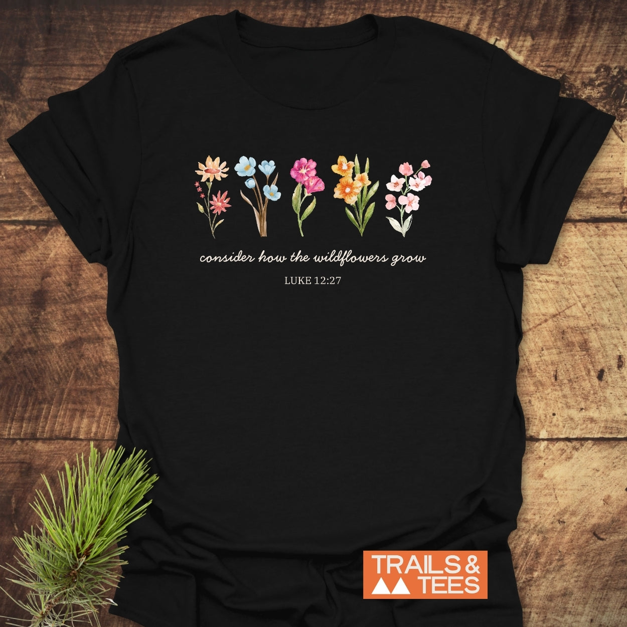 Consider How The Wildflowers Grow, Luke 12:27 T-Shirt features watercolor wildflowers and a quote on black cotton, with a Trails & Tees logo, displayed on a wooden surface with a pine sprig.