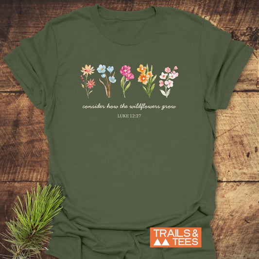 Consider How The Wildflowers Grow, Luke 12:27 T-Shirt, featuring watercolor wildflowers and script text on olive-green fabric, laid flat with a rustic wood backdrop, styled with a pine sprig.