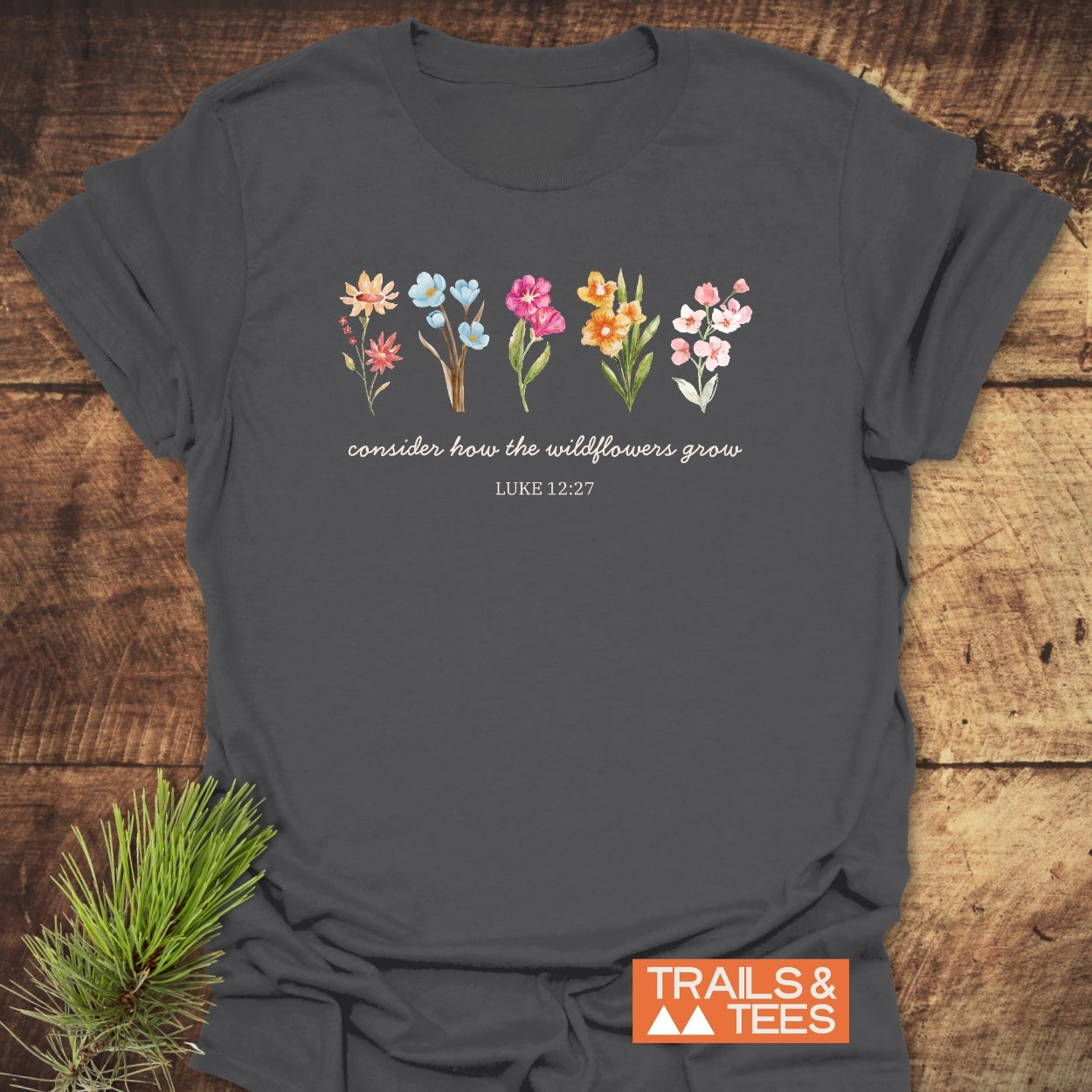 Consider How The Wildflowers Grow, Luke 12:27 T-Shirt features watercolor wildflowers and scripture text on heather-gray cotton, styled with a pine twig, embodying outdoor adventure and comfort.