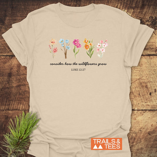 Consider How The Wildflowers Grow, Luke 12:27 T-Shirt, featuring watercolor wildflowers and a script quote, laid flat with TRAILS & TEES logo, showcasing outdoor-inspired design and comfort.