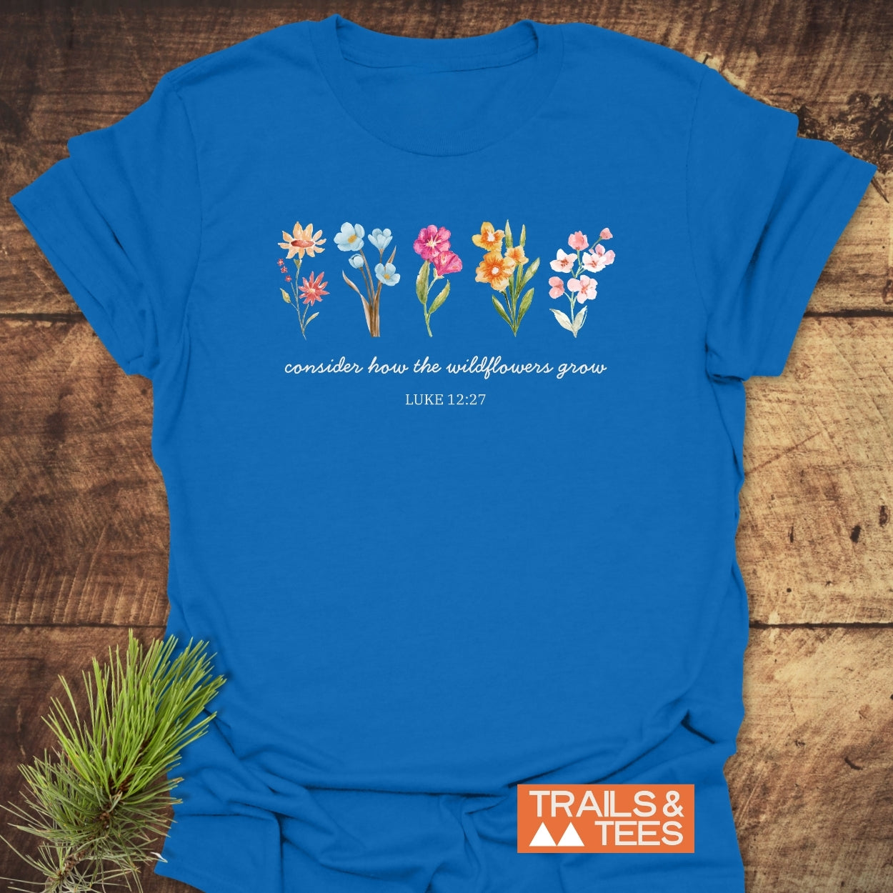 Consider How The Wildflowers Grow, Luke 12:27 T-Shirt featuring watercolor wildflowers and scripture on a cobalt-blue tee, laid on wood with pine greenery, Trails & Tees logo visible.