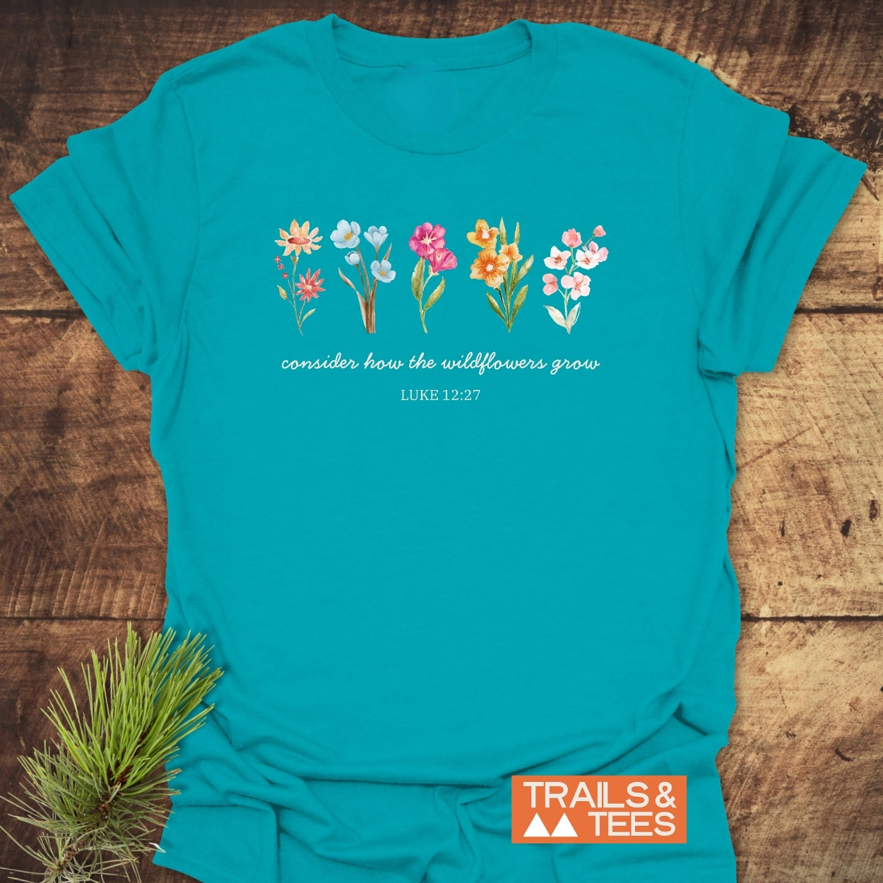 Consider How The Wildflowers Grow, Luke 12:27 T-Shirt features hand-painted wildflowers and script text on teal cotton, laying on wood. Includes TRAILS & TEES logo, symbolizing outdoor adventure and comfort.
