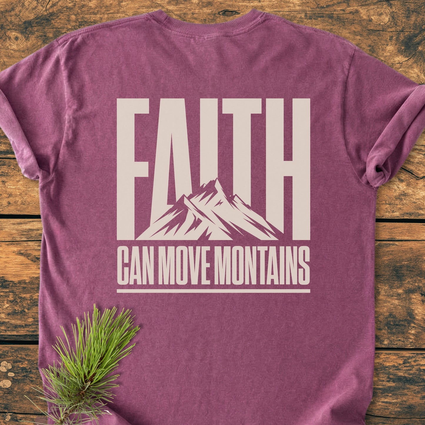 Faith Moves Mountains BACK PRINT - Vintage Wash Tee displays FAITH CAN MOVE MOUNTAINS with a stylized mountain range, laid out flat, styled with a pine branch on a wooden surface.