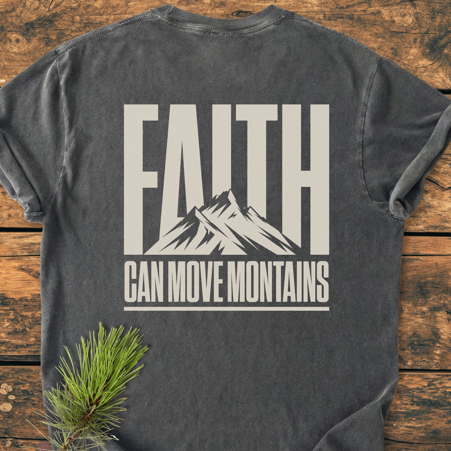 Faith Moves Mountains BACK PRINT - Vintage Wash Tee features a charcoal-gray shade with a bold mountain graphic and inspiring text. Laid on wood, complemented by a pine branch for styling.