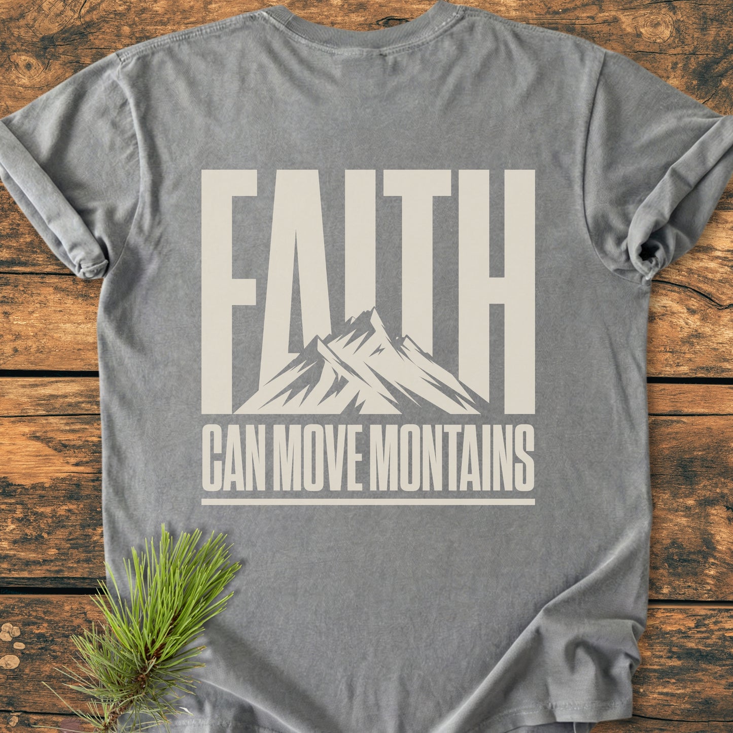 Heather-gray Faith Moves Mountains BACK PRINT - Vintage Wash Tee featuring bold slogan with mountain graphic. Laid on rustic wood, accented by pine sprig, emphasizing nature-inspired theme.
