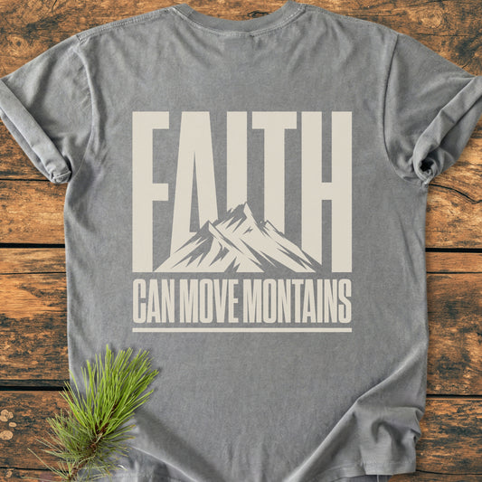 Heather-gray Faith Moves Mountains BACK PRINT - Vintage Wash Tee featuring bold slogan with mountain graphic. Laid on rustic wood, accented by pine sprig, emphasizing nature-inspired theme.