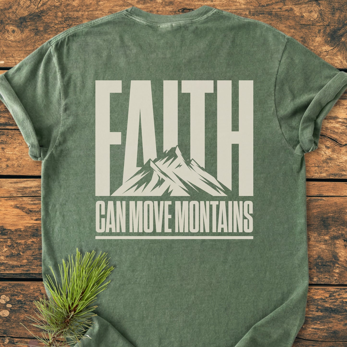 Faith Moves Mountains BACK PRINT - Vintage Wash Tee featuring a bold FAITH with mountain graphic, CAN MOVE MOUNTAINS text beneath, and a pine branch accent on a wooden surface.