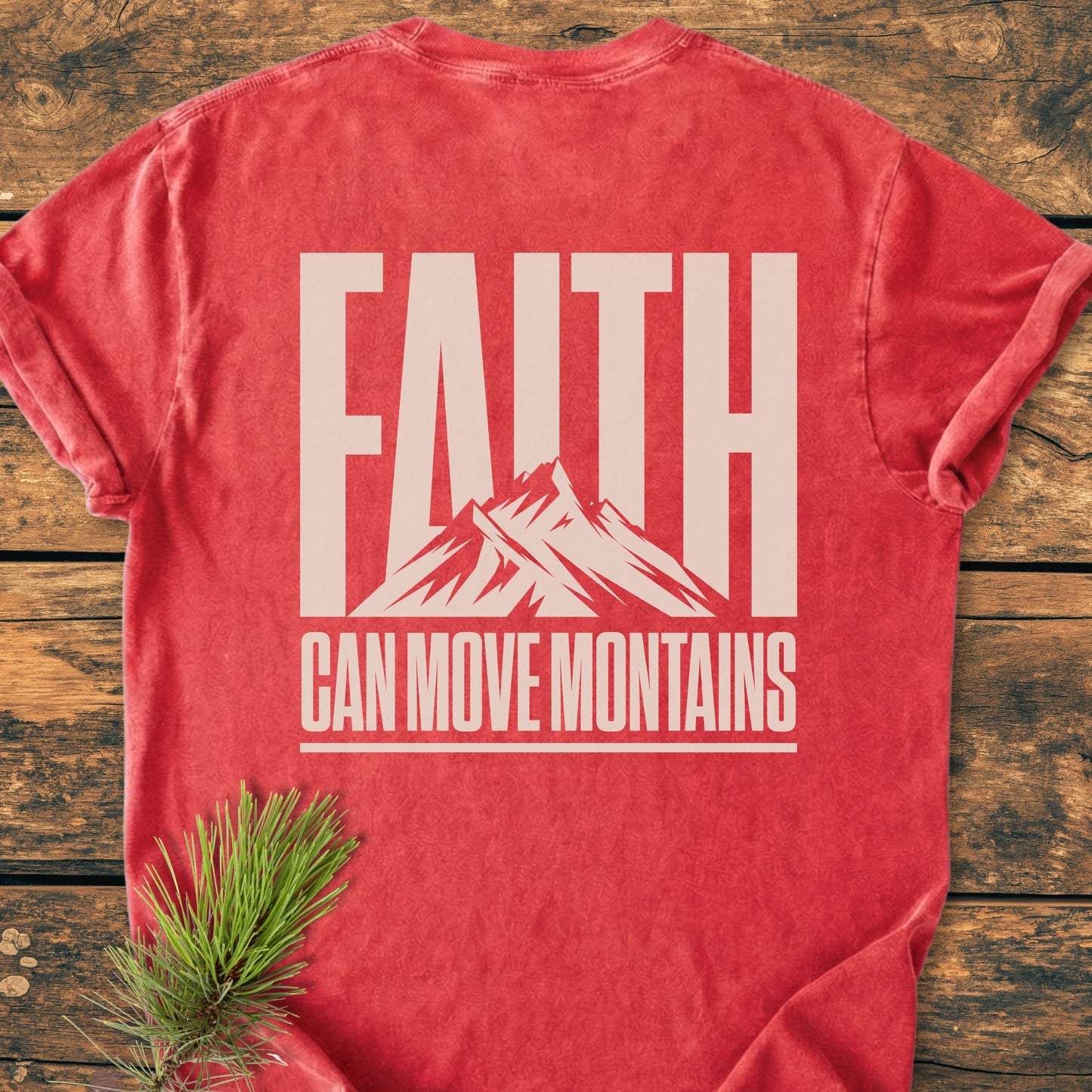 Faith Moves Mountains BACK PRINT - Vintage Wash Tee laid flat on wood. Features 'FAITH' with mountain silhouette, 'CAN MOVE MOUNTAINS' text, pine sprig accent, showcasing garment-dyed, relaxed fit, 100% cotton.