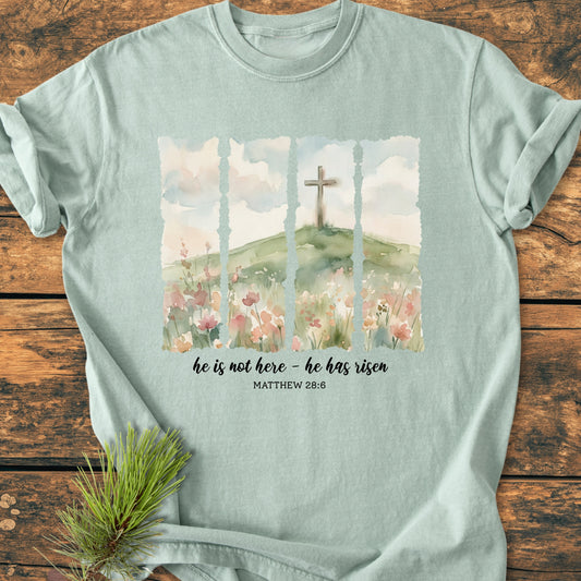 He Is Not Here. He Has Risen - Vintage Heavyweight Tee: Pale mint-green tee featuring watercolor panels of a meadow, hill, and cross, with scriptural caption from Matthew 28:6. Laid on wooden surface with pine sprig.