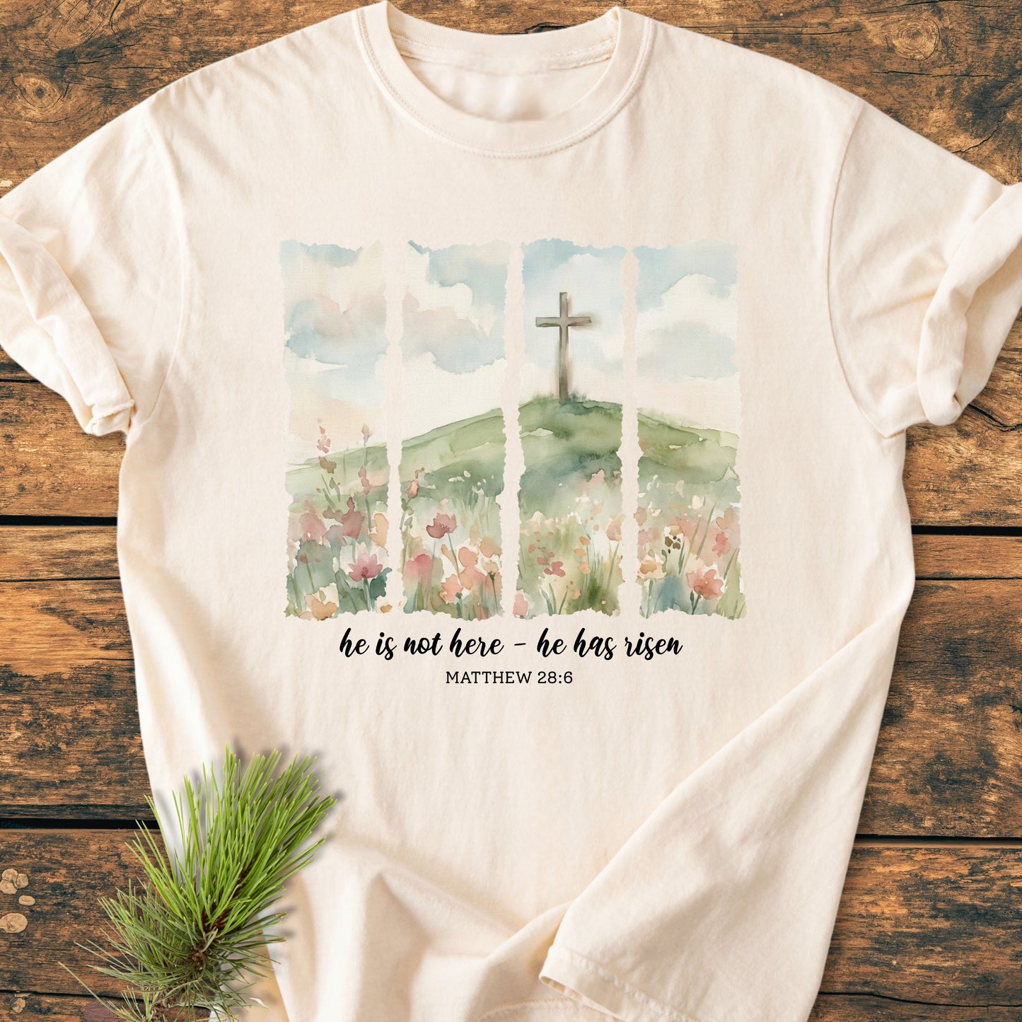 He Is Not Here. He Has Risen - Vintage Heavyweight Tee features a watercolor landscape with a cross and Matthew 28:6 quote, laid on wood, highlighting its comfort and durability.