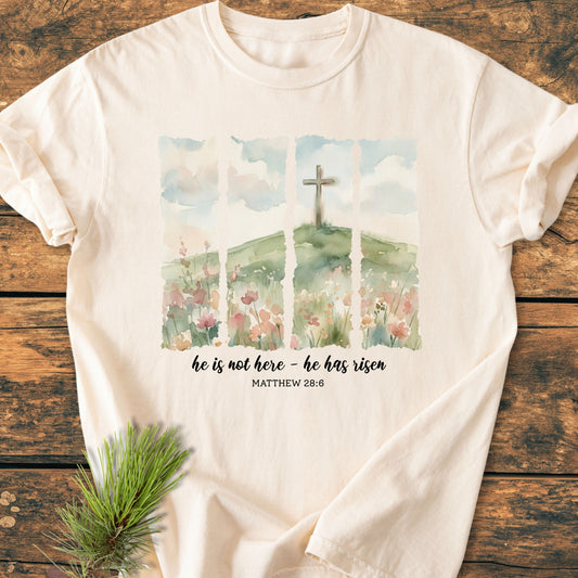 He Is Not Here. He Has Risen - Vintage Heavyweight Tee features a watercolor landscape with a cross and Matthew 28:6 quote, laid on wood, highlighting its comfort and durability.