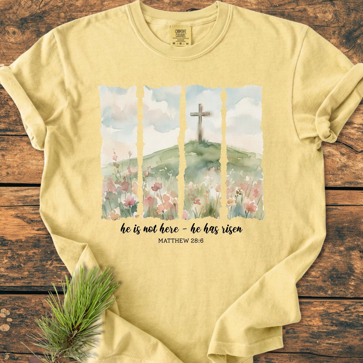 He Is Not Here. He Has Risen - Vintage Heavyweight Tee features a watercolor scene with a cross, wildflowers, and Bible verse from Matthew 28:6 on a pale-yellow, relaxed fit, garment-dyed t-shirt.