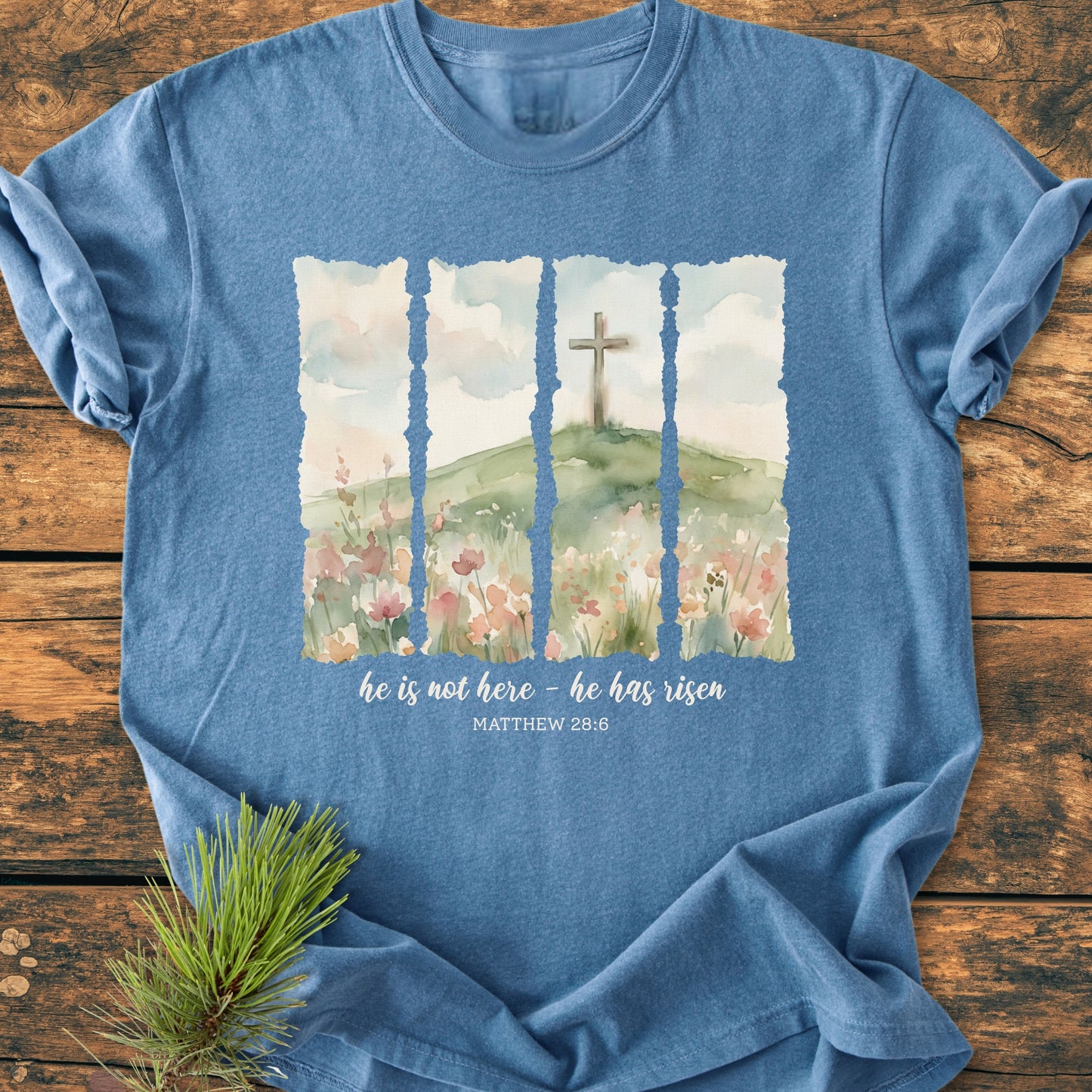 He Is Not Here. He Has Risen - Vintage Heavyweight Tee features a watercolor hill scene with a cross and scripture text, laid flat on wood, emphasizing its relaxed fit and durability.