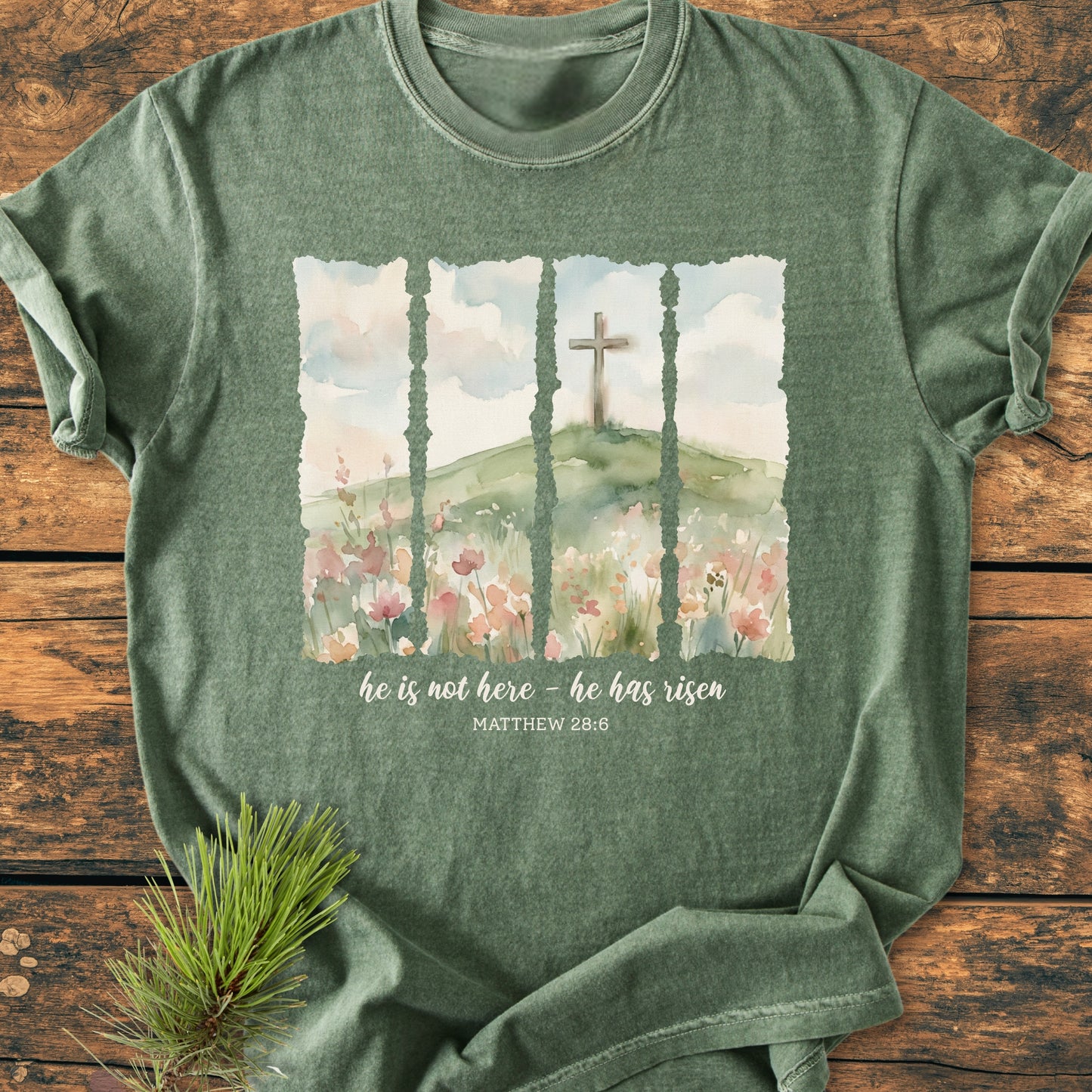 He Is Not Here. He Has Risen - Vintage Heavyweight Tee featuring a watercolor-style design with a hill, wildflowers, and cross, laid flat on rustic wood, highlighting comfort and durability.