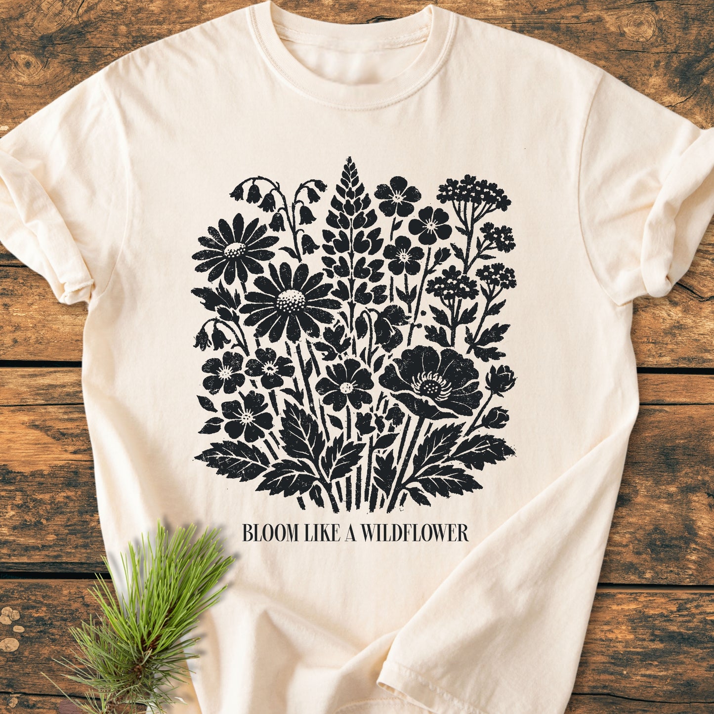 Bloom Like A Wildflower - Vintage Heavyweight Tee features a black wildflower print on a light-cream T-shirt, laid on a wooden surface with a pine sprig beside it.