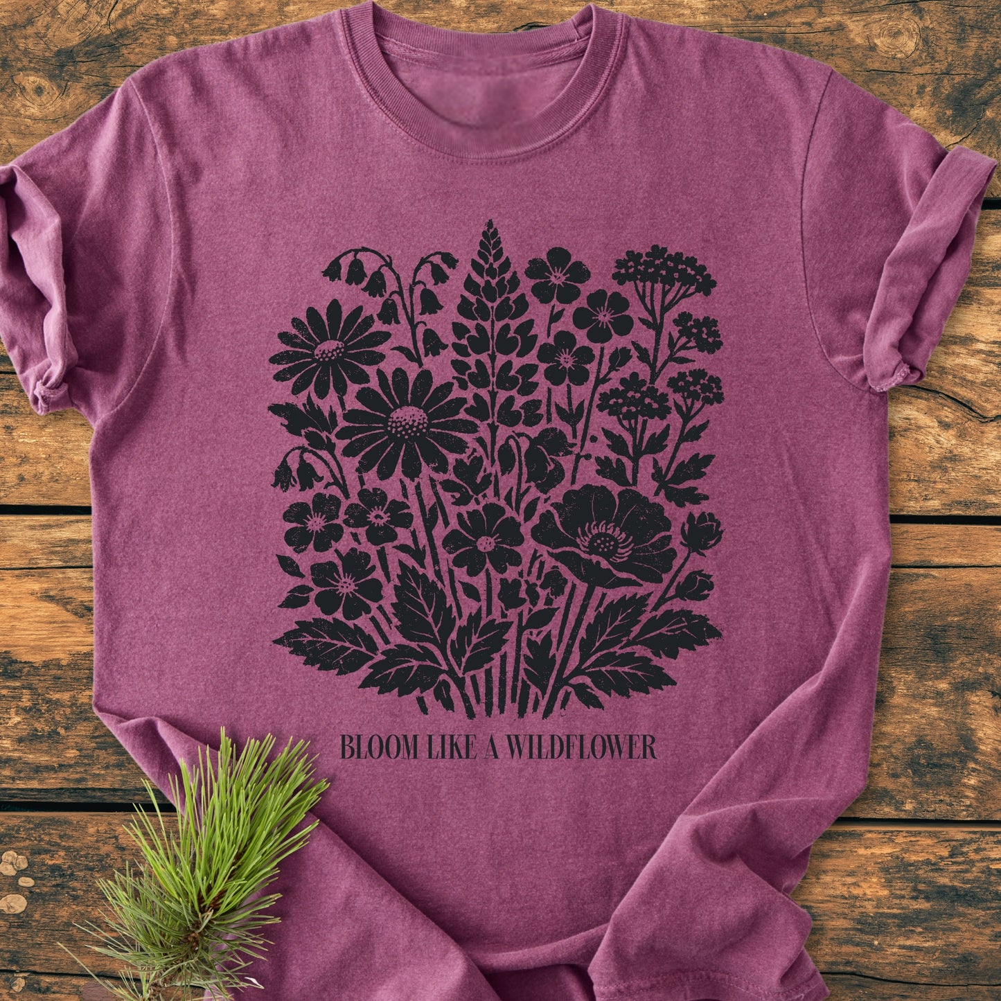 The Bloom Like A Wildflower - Vintage Heavyweight Tee showcases a heather-purple color with black wildflower print and slogan, styled on rustic wood with a pine sprig accent.