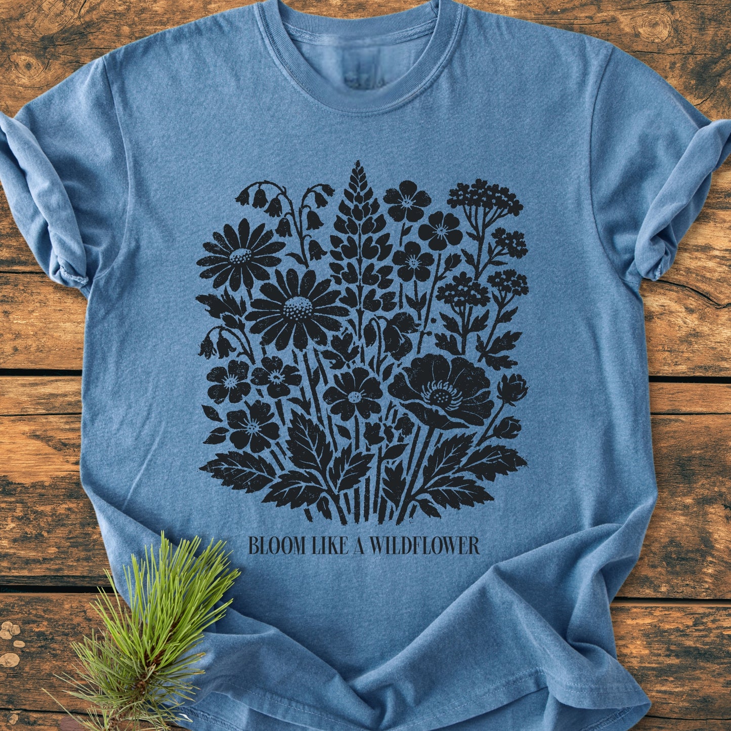 Heather-blue Bloom Like A Wildflower - Vintage Heavyweight Tee featuring a black wildflower design and slogan, laid flat on wooden surface. A sprig of pine accents the scene.