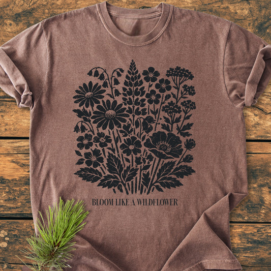 The Bloom Like A Wildflower - Vintage Heavyweight Tee features wildflower silhouettes and a slogan on a brown heathered T-shirt, styled on a rustic wooden surface with a pine sprig accent.