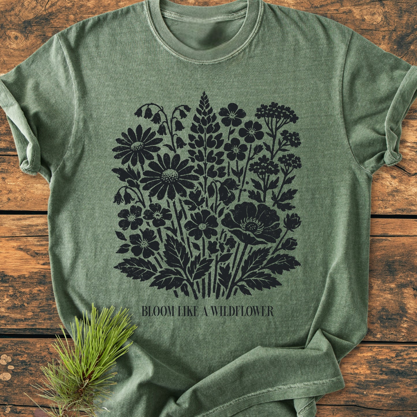 Bloom Like A Wildflower - Vintage Heavyweight Tee features a heather-green design with a black wildflower silhouette and text. Displayed on wood with a pine garnish, highlighting its nature-inspired theme.
